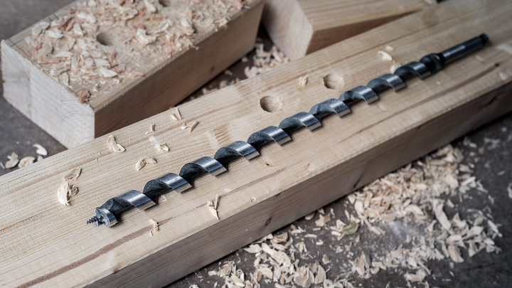 Object #200461326: 3088579_app_web_2 Large auger drill bit resting on a wooden beam with wood shavings scattered around.